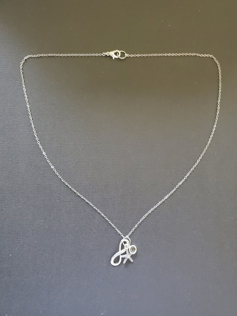 May include: A silver-colored necklace with a delicate chain and a pendant. The pendant features a stylized letter "A" intertwined with a small figure, possibly representing a person or a symbol of connection. The necklace is displayed against a dark background.