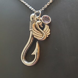 May include: Silver-toned necklace featuring a large fish hook pendant with a swan charm and a small, round, purple gemstone. The chain is delicate, and the pendant has intricate details. The necklace is displayed against a dark background.