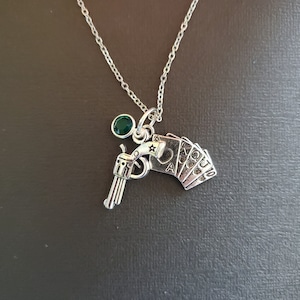 Peut inclure: Collier argenté avec un pendentif revolver et un éventail de cartes à jouer. Une petite pierre verte est également attachée. Le collier est présenté sur un fond sombre.