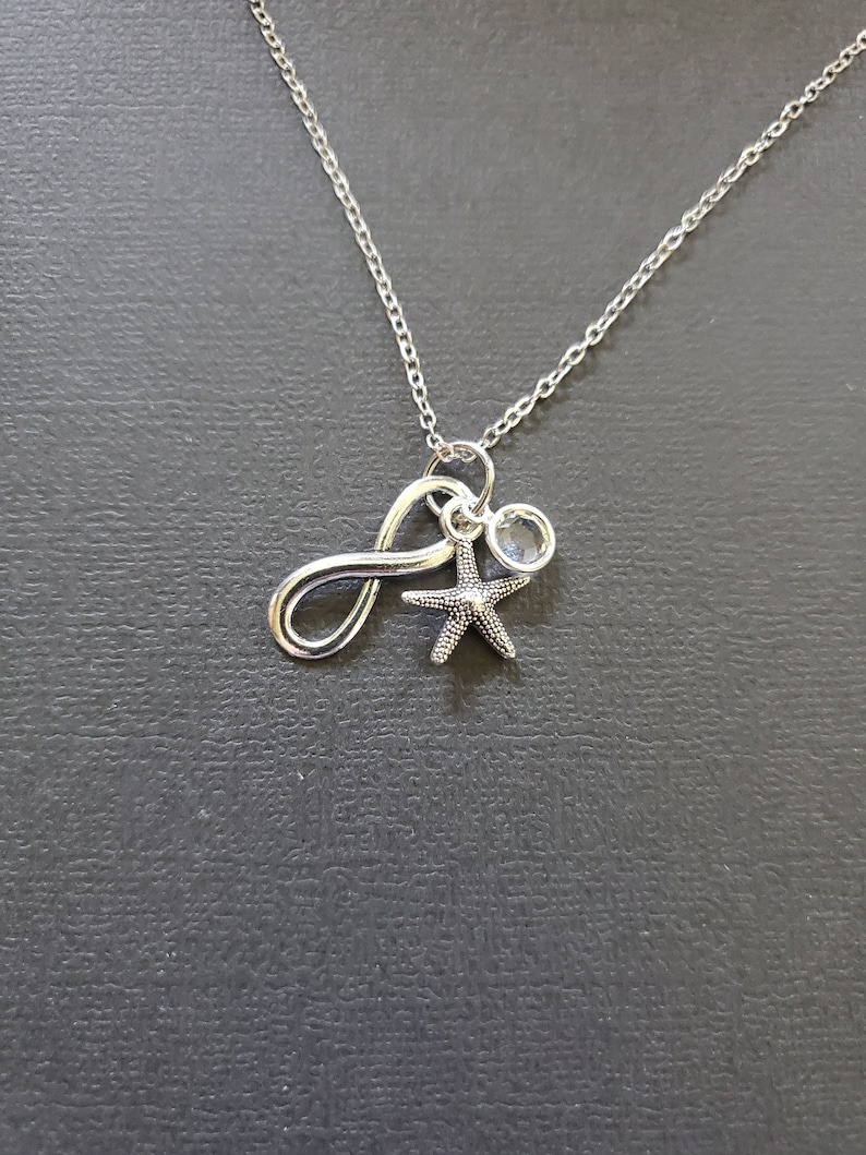 May include: Silver-toned necklace with a delicate chain. The pendant features an infinity symbol, a small starfish charm, and a clear, round crystal. The necklace is displayed against a dark gray background.