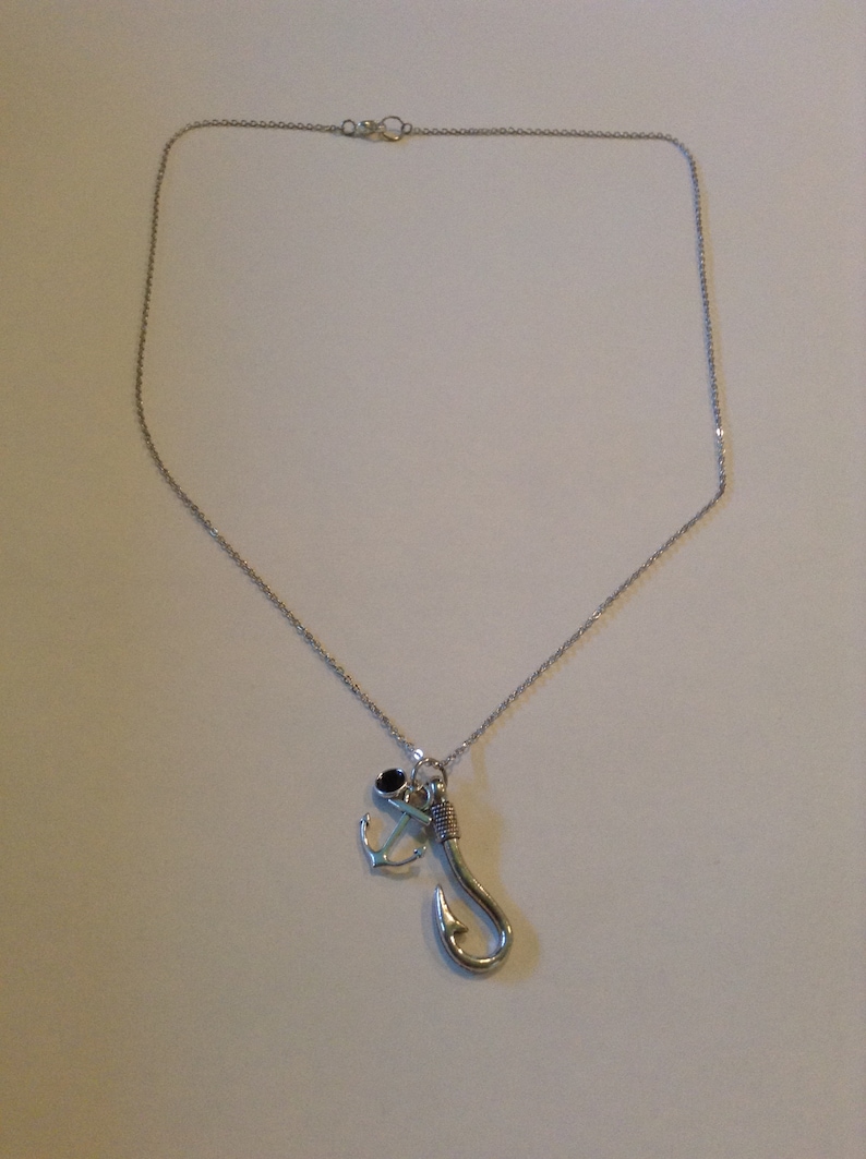 May include: Silver-toned necklace featuring a fishing hook and anchor pendant. The hook is detailed with wrapped wire, and the anchor has a small black accent. The chain is delicate, and the clasp is visible at the top.