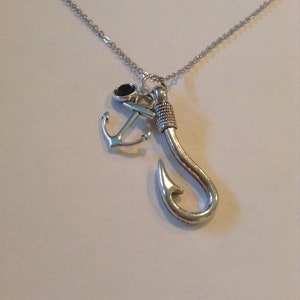 May include: Silver-toned necklace featuring a fishing hook pendant, an anchor charm, and a small black stone. The chain is delicate, and the pendants are detailed. A nautical-themed jewelry piece.