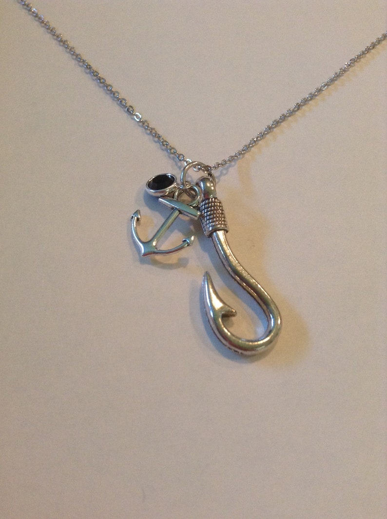May include: Silver-toned necklace featuring a fishing hook pendant, an anchor charm, and a small black stone. The chain is delicate, and the pendants are detailed. A nautical-themed jewelry piece.