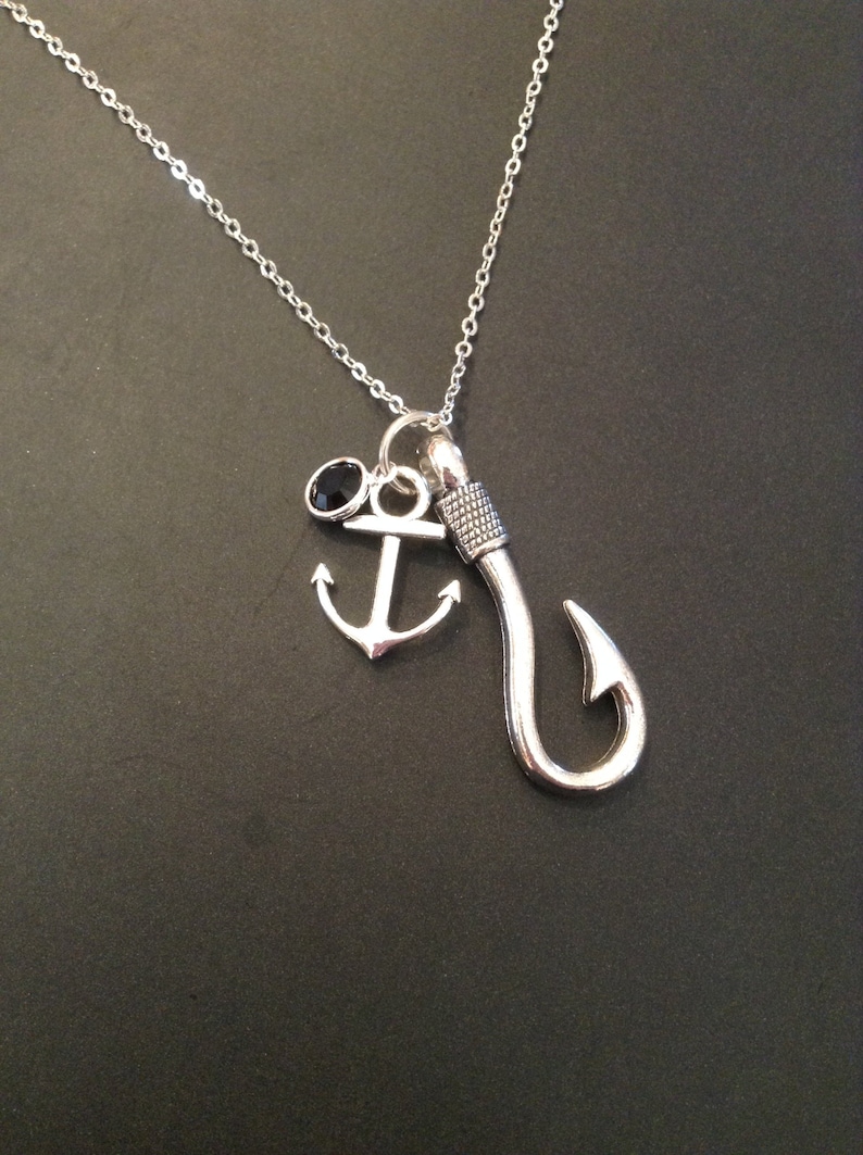 May include: Silver-toned necklace featuring a fishing hook charm, an anchor charm, and a small black gemstone. The chain is delicate, and the charms are detailed. The necklace is displayed against a dark background.