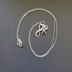 May include: A silver necklace with a delicate chain. The pendant features the letter "J", a starfish, and a small clear crystal. The necklace is displayed on a dark gray surface.