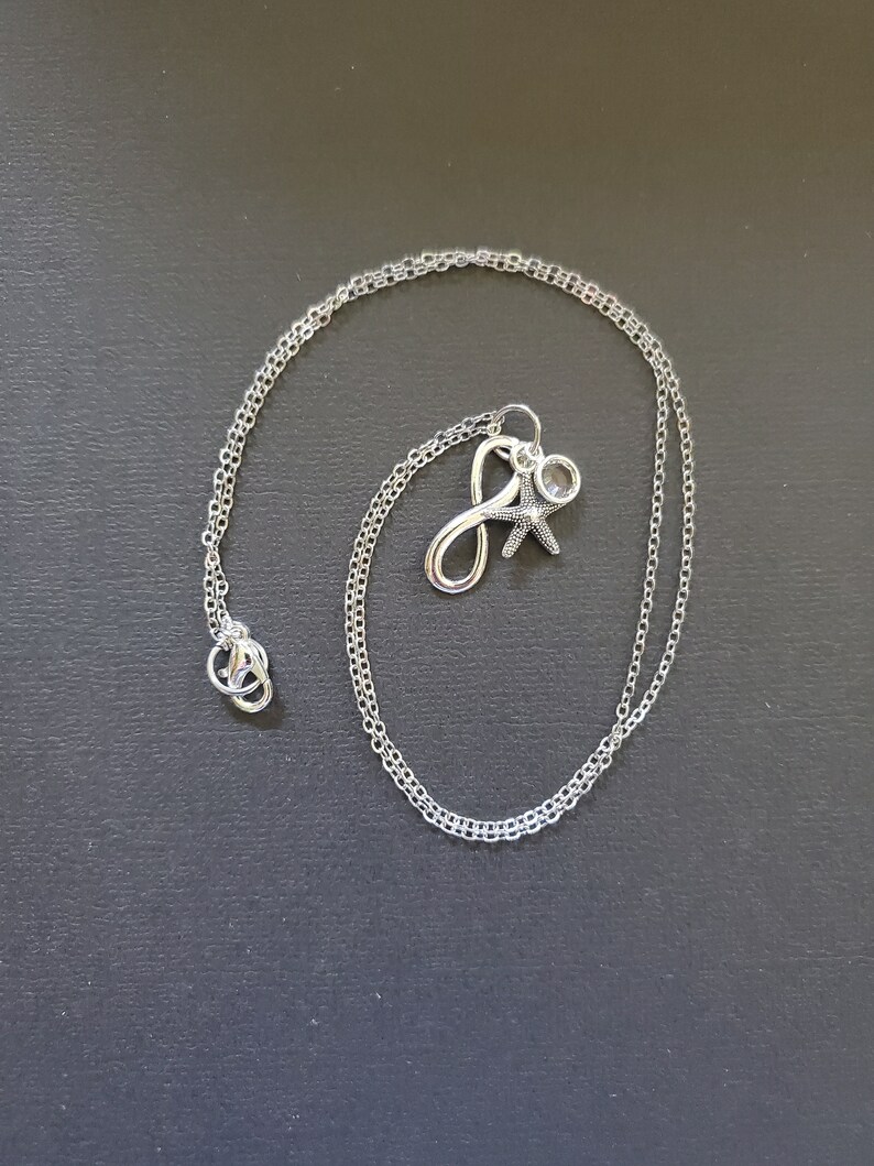 May include: A silver necklace with a delicate chain. The pendant features the letter "J", a starfish, and a small clear crystal. The necklace is displayed on a dark gray surface.