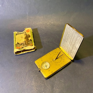 May include: A vintage gold-toned metal Sunwatch pocket sundial and compass with a hinged lid featuring a printed table of data. The device includes a central gnomon and a small circular compass. It is accompanied by a worn, illustrated cardboard box.