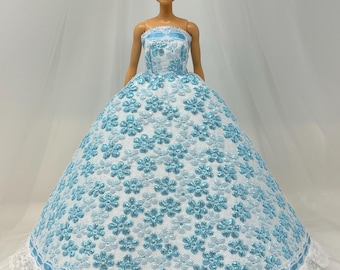 May include: A doll wearing a strapless, light blue and white floral patterned gown with a white lace trim. The doll has blonde hair styled in a bun. The dress has a full skirt and a fitted bodice.