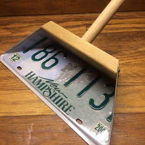 May include: A wooden dustpan made from a repurposed New Hampshire license plate with the number 186113. The license plate is silver with green lettering and a picture of a mountain range. The wooden handle is attached to the top of the license plate.