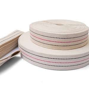 May include: Two rolls of off-white cotton webbing with black and red accent stripes. A separate length of the same webbing is visible to the left. The webbing is tightly wound and appears to be new.