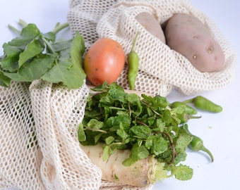 May include: A white mesh produce bag filled with fresh vegetables, including green leafy vegetables, a red tomato, green peppers, and a white radish. The bag is sitting on a white surface.
