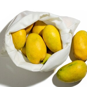 May include: A white reusable produce bag filled with yellow mangoes. Two additional mangoes are sitting outside the bag on a white surface.