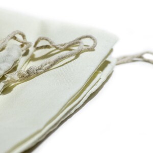 May include: A white cotton drawstring bag with a natural cotton cord. The bag is folded and the cord is tied in a knot.