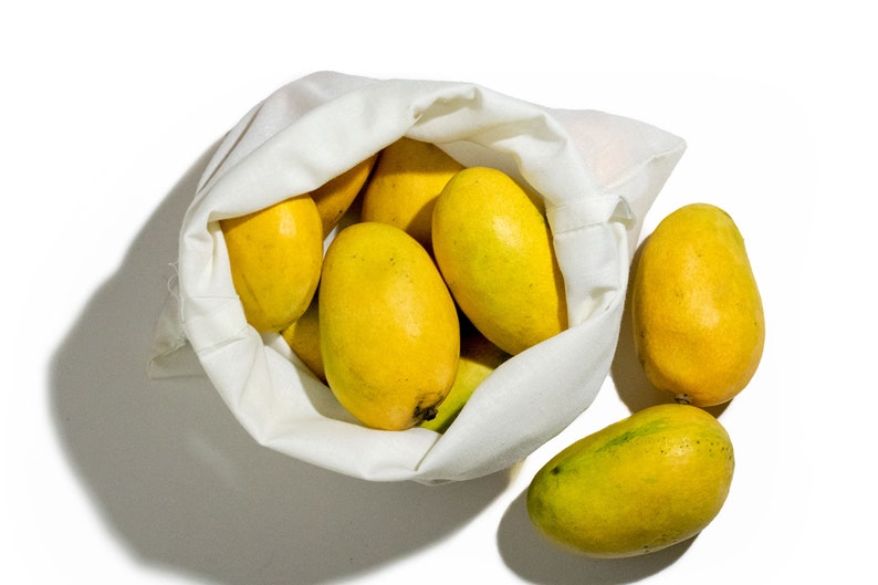 May include: A white reusable produce bag filled with yellow mangoes. Two additional mangoes are sitting outside the bag on a white surface.