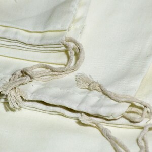 May include: A stack of white cotton drawstring bags with natural cotton drawstrings. The bags are folded and stacked on top of each other.