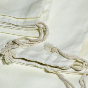 May include: A stack of white cotton drawstring bags with natural cotton drawstrings. The bags are folded and stacked on top of each other.