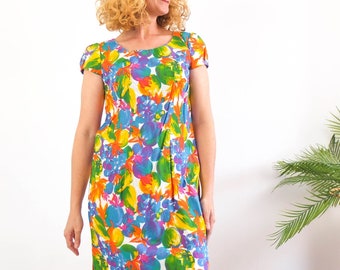 Puede incluir: Una mujer lleva un vestido de manga corta con estampado floral de colores. El vestido tiene un escote redondo y una silueta ajustada. La mujer está de pie frente a una pared blanca.