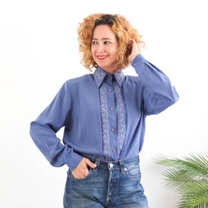 May include: A periwinkle blue long-sleeved blouse with a collared neckline and button-down front. The blouse has delicate white embroidery on the collar and front. The model is wearing blue jeans.