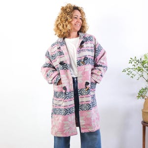 May include: A long, patterned coat in shades of pink, gray, and navy blue. The coat features a geometric design and wooden toggle closures. The coat is worn over a white shirt and blue jeans.