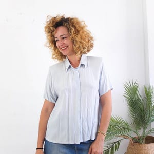 Pode incluir: Camisa azul clara de manga curta com um padrão xadrez subtil. A camisa tem uma gola clássica e fecho de botões. É uma peça de vestuário vintage, feita de tecido leve, ideal para o verão.