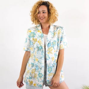 Peut inclure: Un blazer blanc à motifs floraux dans les tons de bleu et de jaune. Le blazer a des manches courtes et un col cranté. Le mot "Sister" est imprimé sur le blazer.