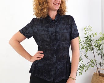 May include: A dark blue and grey patterned short-sleeved shirt with a button-down front and a collared neckline. The shirt is fitted at the waist. The model is wearing black trousers and a gold bracelet.