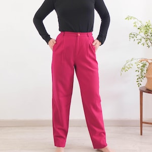 May include: A person wearing a black roll-neck top and high-waisted, magenta trousers. The trousers have a button closure and pleats at the waist. The outfit is simple and classic.