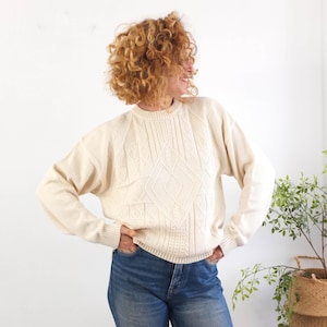 Könnte beinhalten: Cremefarbener Pullover mit Rauten- und Zopfmuster. Der langärmlige Pullover hat einen Rundhalsausschnitt und gerippte Bündchen und Saum. Der Pullover wird mit einer blauen Jeans getragen. Die Person lächelt und hat lockiges Haar.