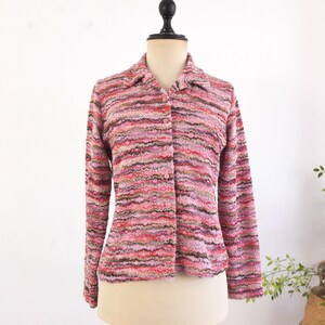 Op de afbeelding: Een roze, veelkleurig gebreid vest met lange mouwen en een kraag. De trui met knopen heeft tinten roze, rood en bruin. Het kledingstuk wordt op een mannequin getoond.