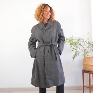 Peut inclure: Un trench-coat gris avec une taille ceinturée et un col classique. Le manteau arrive au genou et a des manches longues. La personne porte un pantalon noir et des bottes. Le manteau est en tissu texturé.