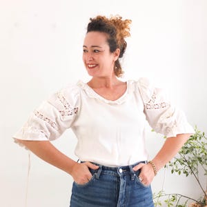 May include: White blouse with a scalloped neckline and lace detailing on the sleeves. The blouse has a relaxed fit with three-quarter sleeves and is paired with blue denim jeans. The overall style is casual and feminine.