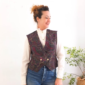 Può includere: Un gilet con stampa paisley nei toni del bordeaux, oro e blu navy. Il gilet ha un design doppiopetto con bottoni dorati. È indossato sopra una camicia bianca con colletto e jeans blu.