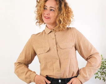 May include: A tan and white checked shirt with two front pockets. The long-sleeved shirt has a collared neckline and is worn with black trousers. The shirt is made of a lightweight fabric.