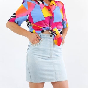 May include: A woman wearing a short-sleeved, multi-colored button-up shirt tied at the waist and a light blue denim skirt with a floral trim.