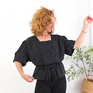 May include: Black short-sleeved top with a square neckline and a tie waist. The top has a subtle geometric pattern and is paired with black pants. The model is wearing a gold bracelet. A plant in a woven basket is in the background.