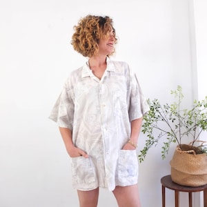 Pode incluir: Uma camisa de manga curta de cor clara com um padrão subtil e repetitivo. A camisa tem gola, fecho de botões na frente e dois bolsos frontais grandes. A peça de roupa é folgada e parece ser feita de um tecido leve.