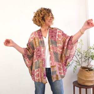 May include: A patchwork kimono-style jacket in shades of pink, gold, and gray. The open-front garment features a paisley pattern and is worn over a white top and blue jeans. The sleeves are wide and flowing.