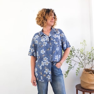 Pode incluir: Uma camisa azul com botões e padrão floral. A camisa tem mangas curtas e dois bolsos no peito. O design floral inclui flores brancas e azul claro com caules bege. A camisa é combinada com jeans azuis.