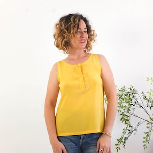 May include: A sleeveless bright yellow blouse featuring a round neckline and a central vertical placket detail with a single matching button. The top is paired with blue denim jeans, creating a simple and clean look for everyday wear.