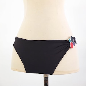 May include: Black bikini bottoms with a decorative flower detail on the side. The flower is made of overlapping oval shapes in red, blue, green, and purple. The bikini bottoms are displayed on a mannequin.