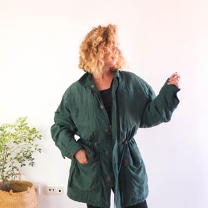 Può includere: Una giacca verde scuro con colletto, maniche lunghe e chiusura frontale con bottoni. La giacca presenta due ampie tasche anteriori e un cordoncino in vita, offrendo una vestibilità strutturata con una finitura in tessuto opaco.