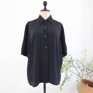 May include: A black button-down shirt with short sleeves, displayed on a mannequin. The shirt features a collared neckline with delicate embroidered detailing and a series of buttons down the front. The fabric appears to be lightweight and slightly sheer.