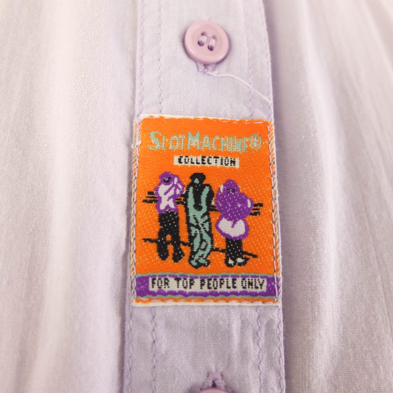 May include: Close-up of a light purple button-down shirt. An orange label with the text "SHOT MACHINE COLLECTION" and three cartoon figures is sewn onto the shirt. The text "FOR TOP PEOPLE ONLY" is also on the label.