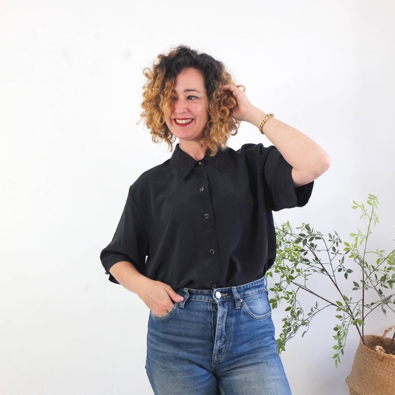 May include: Black button-down shirt with short sleeves and a decorative collar. The shirt is paired with blue jeans. A woman is wearing the shirt and jeans. A plant in a woven basket is in the background.