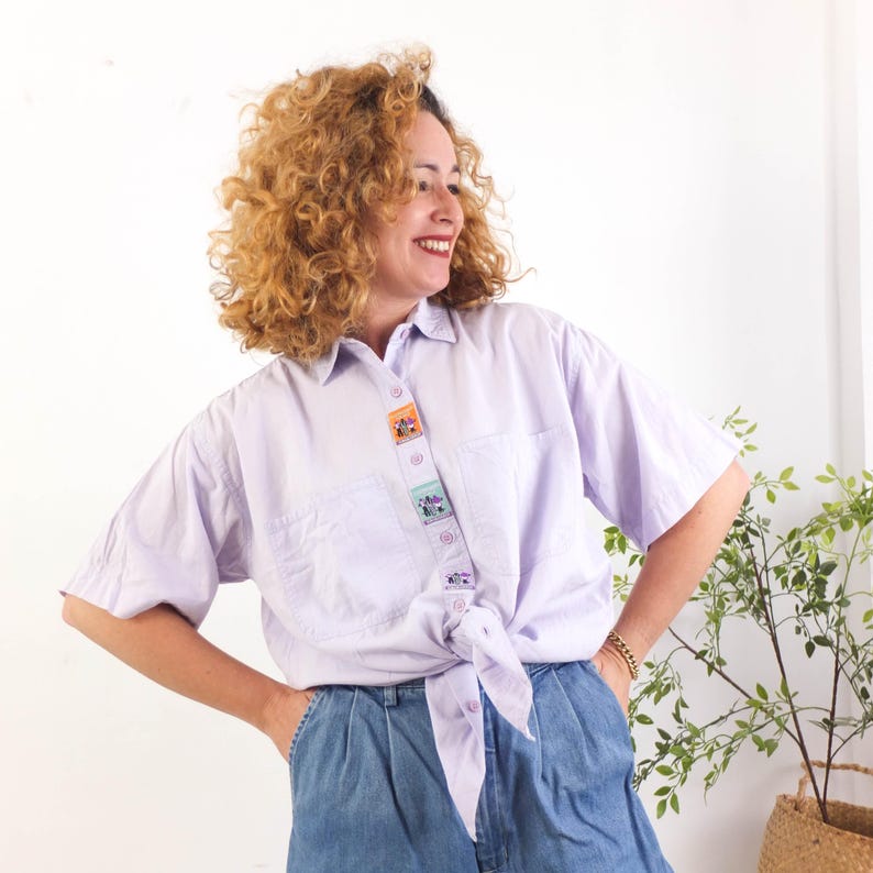 May include: A light purple button-down shirt with short sleeves and a tied knot at the waist. The shirt features a vertical strip of colorful, decorative patches down the front. The shirt is paired with blue denim shorts.