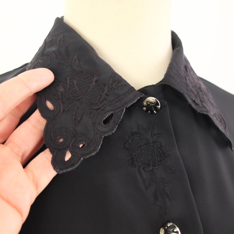 May include: Close-up of a black blouse featuring an embroidered collar with a scalloped edge. The collar and cuffs have floral embroidery. The blouse has decorative buttons.