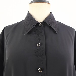 May include: A black button-down shirt with a decorative collar and floral embroidery down the front. The collar has a scalloped edge. The buttons are silver-toned. The shirt is displayed on a mannequin.