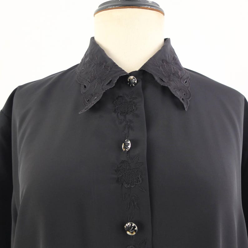 May include: A black button-down shirt with a decorative collar and floral embroidery down the front. The collar has a scalloped edge. The buttons are silver-toned. The shirt is displayed on a mannequin.