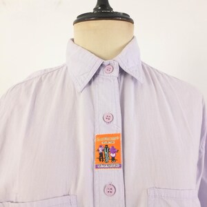 May include: A light purple button-down shirt with a collar and two chest pockets. The shirt features a rectangular patch with cartoon figures and the text "ScaMachina Collection" and "WE LOVE PEOPLE SHOP".