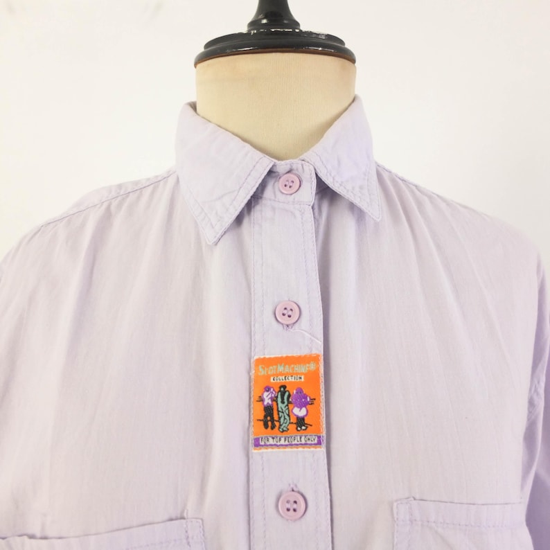 May include: A light purple button-down shirt with a collar and two chest pockets. The shirt features a rectangular patch with cartoon figures and the text "ScaMachina Collection" and "WE LOVE PEOPLE SHOP".